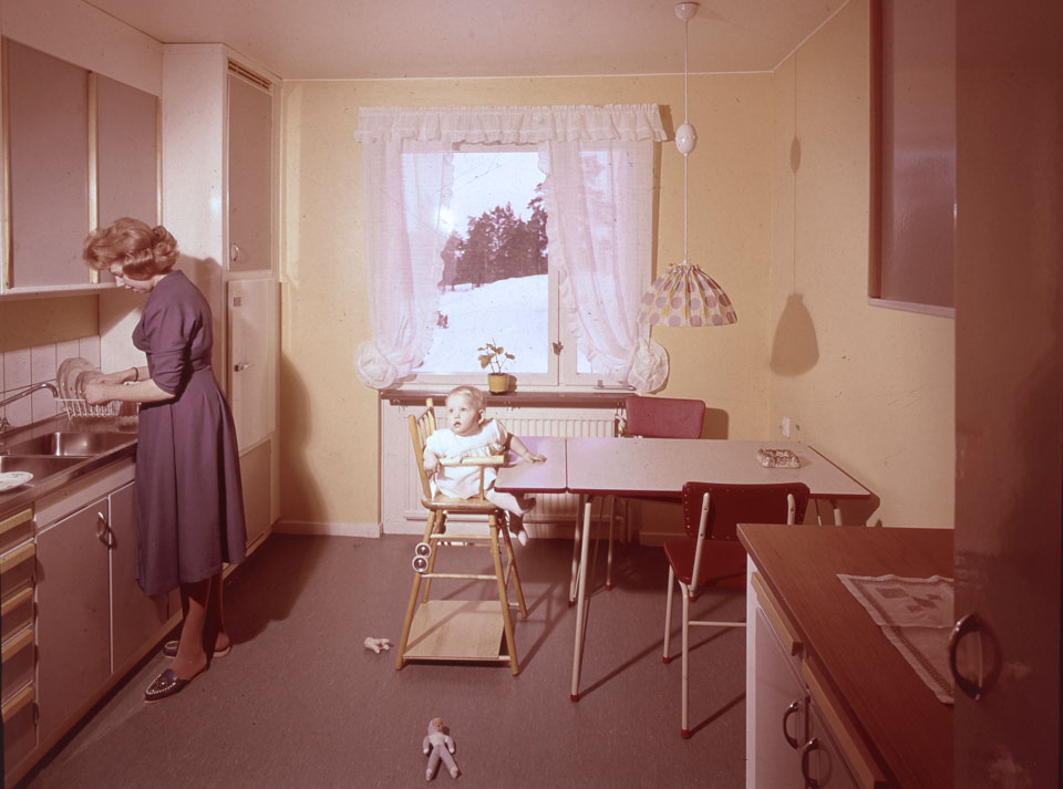 HSB-kök från 50-talet. En kvinna står och arbetar vid köksbänken. Vid matbordet sitter ett barn i en barnstol.