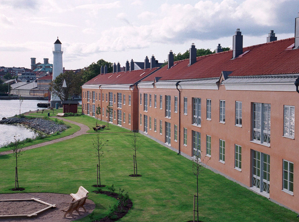 Foto på fasaden av Stumholmen, byggnaden har röd puts och röda takpannor. Bredvid huset finns vatten och en fyr.