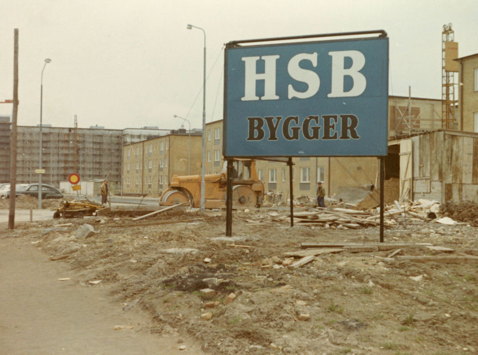 En blå skylt med texten "HSB bygger" framför en byggarbetsplats där nya bostäder håller på att byggas.