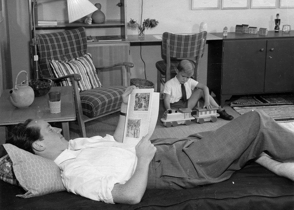En man liggandes på en soffa som läser en tidning och en pojke på golvet som leker